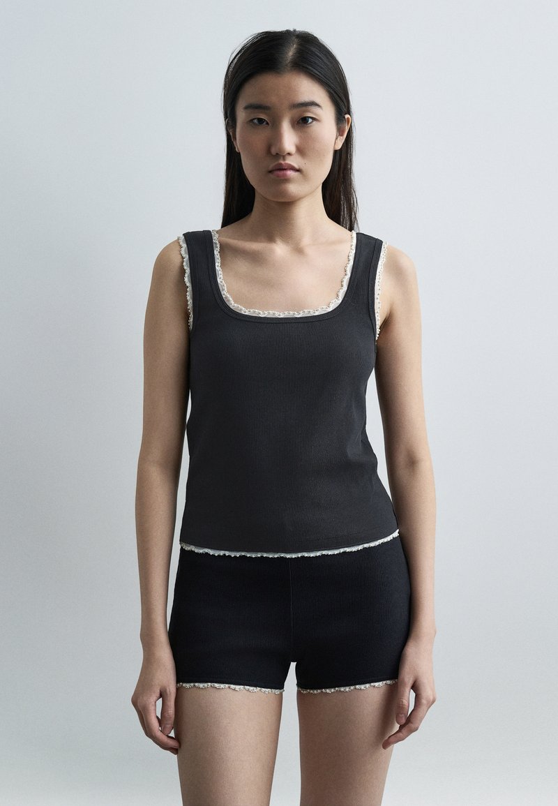 Femme aux longs cheveux bruns portant un haut noir sans manches et un short assorti avec une bordure de dentelle blanche, debout devant un fond gris uni.