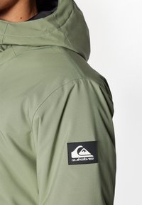 Zöld vízálló dzseki kapucnival. Fekete Quiksilver logó címkével a ruhaujján. Simaságot és varrási részleteket mutat.