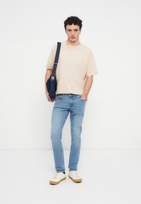 Μπεζ κοντομάνικο μπλουζάκι συνδυασμένο με ανοιχτό μπλε τζιν, κρατώντας μια σκούρου μπλε τσάντα ώμου. Τα αθλητικά παπούτσια έχουν εκρού επάνω μέρος και καφέ σόλα.