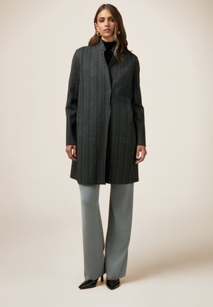 Cappotto lungo grigio carbone con taglio dritto e motivo testurizzato, indossato sopra pantaloni grigi aderenti e décolleté neri a punta.