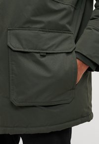 Chaqueta verde acolchada con un acabado texturizado, que presenta un gran bolsillo en ángulo en la parte frontal y un puño acanalado en la manga.