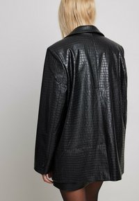 Blonde person wearing an oversized black faux leather blazer with crocodile texture, shown from the back against a plain background.