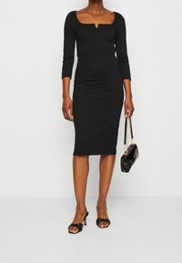 Musta muotoon liittyvä mekko pitkilla hihoilla ja neliönmuotoisella pääntiellä, jossa on etuleikkaus. Yhdistetty mustiin korkokenkiin ja mustaan käsilaukkuun.