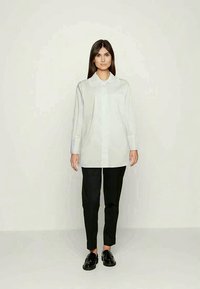 Camisa branca de manga longa com colarinho, combinada com calças pretas ajustadas e sapatos pretos, apresentando tecidos suaves e design minimalista.