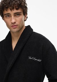 Svart frottébadekåpe med sjalkrage, med hvit brodert "Karl Lagerfeld"-logo på venstre side. Myk tekstur, knelengde.