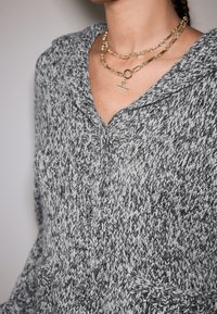 Pletena dukserica s kapuljačom u sivoj i bijeloj boji s teksturiranim uzorkom, s V-izrezom, gumbima i dva džepa. Vidljive zlatne slojevite lančane ogrlice.