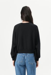 Maglione nero corto con maniche raglan, caratterizzato da dettagli frangiati sui lati. Abbinato a jeans in denim blu a vita alta.