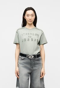 Γυναίκα που στέκεται και κοιτά μπροστά, φορώντας μια φθαρμένη πράσινη μπλούζα με τυπωμένο κείμενο και ψηλόμεσα ξεβαμμένα γκρι τζιν με μαύρη ζώνη.