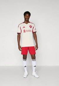 Άνδρας που φοράει λευκή και κόκκινη φανέλα ποδοσφαίρου της Λίβερπουλ με αντίστοιχες κάλτσες και παπούτσια ποδοσφαίρου, στέκεται μπροστά σε απλό ανοιχτό φόντο.