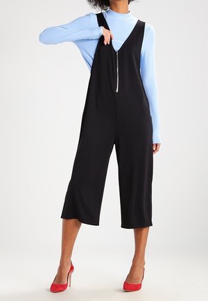 Zwarte overall jumpsuit met een ritssluiting aan de voorkant, wijde pijpen en een relaxte pasvorm, gecombineerd met een lichtblauwe blouse met lange mouwen en rode hakken.
