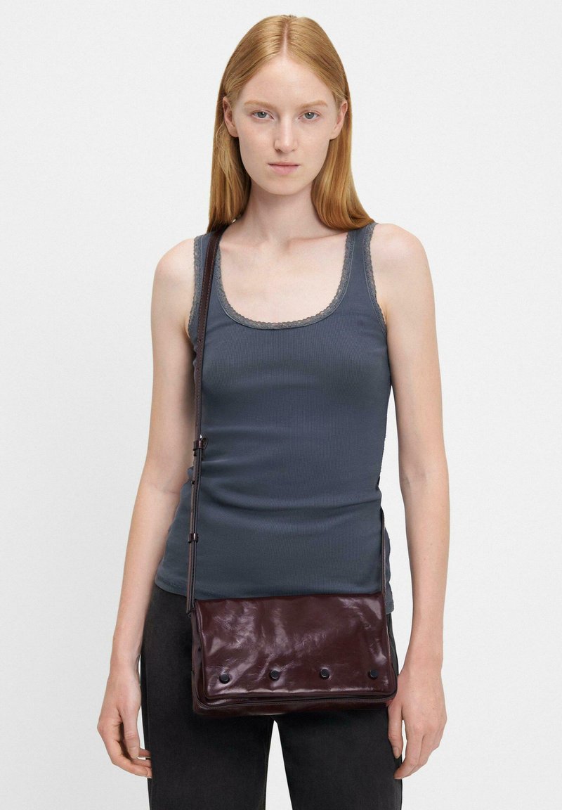 Una borsa a tracolla in pelle bordeaux scuro con una texture liscia, caratterizzata da una patta anteriore e cinque dettagli a pulsante. Indossata sulla spalla di un modello.