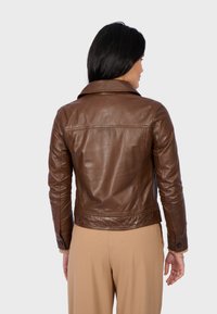 Veste en cuir marron avec un col, au design ajusté et des détails de couture visibles. Portée sur un pantalon beige, vue de dos.