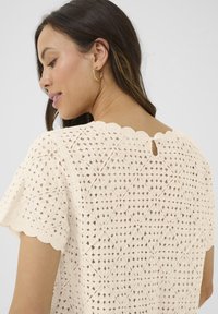 Top in pizzo color crema caratterizzato da un collo a scallop, un motivo a lavorazione aperta e maniche corte a volants con chiusura a bottone sul retro.