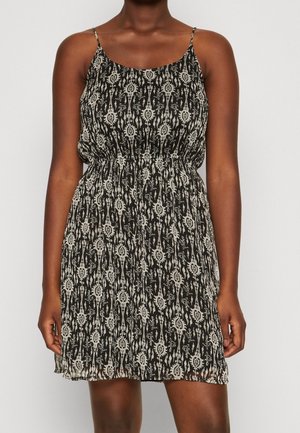 Kobieta w czarnej sukience bez rękawów z białym abstrakcyjnym kwiatowym wzorem i talią podkreśloną paskiem, stojąca na jednolitym tle.