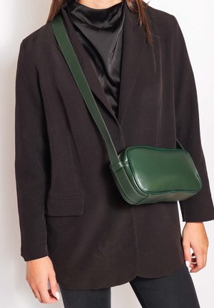 Persona che indossa una camicetta di raso nera e un blazer con una borsa a tracolla in pelle verde scuro con una tracolla larga attraversata sul petto.