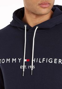 Felpa blu navy con cordini bianchi, caratterizzata da testo e logo ricamati "TOMMY HILFIGER" in rosso e bianco. Tessuto morbido.