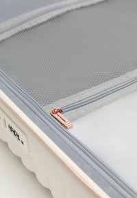 Light-colored hard-shell suitcase with a mesh zippered compartment and a combination lock on the side.