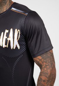 Zwarte sportshirt met een stippenpatroon, voorzien van een groot wit logo en oranje accenten. Textuurstof, korte mouwen en een ronde hals.