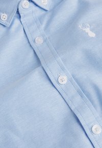 Chemise bleu clair boutonnée avec des boutons blancs et un petit logo brodé blanc représentant une tête de cerf sur la poitrine.