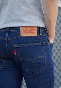 Jeans de ganga azul com um acabamento texturizado, apresentando uma etiqueta de couro, costura amarela e um rótulo vermelho no bolso traseiro. Design de ajuste padrão.