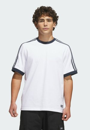 Jeune homme aux cheveux bouclés portant un t-shirt blanc avec des rayures bleu marine sur les épaules et les manches, debout devant un fond uni.