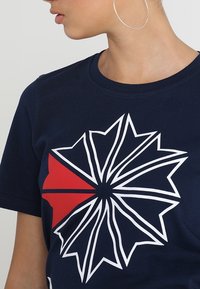 Námořnická modrá tričko s grafickým designem, které obsahuje bílý vzor slunečního paprsku a červený akcent, vyrobené z bavlny s kulatým výstřihem.