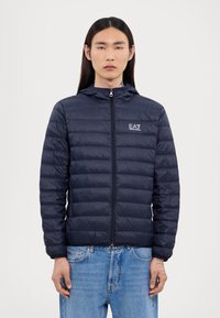 Doudoune navy avec fermeture éclair sur le devant et capuche, présentant un design matelassé et le logo EA7 sur la poitrine. Portée avec un jean bleu clair.
