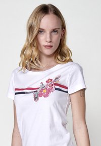 Camiseta blanca de algodón con mangas cortas, con rayas en rosa y azul y bordado floral. Texto "KOROSHI" encima del diseño.
