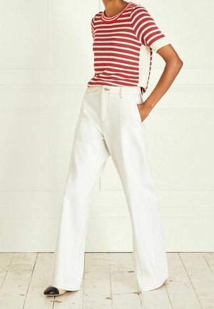 Person trägt ein rot-weiß gestreiftes, kurzärmeliges Strickoberteil, das in hoch taillierte, weitbeinige weiße Hosen gesteckt ist, dazu schwarze Schuhe mit Spitzen in Schwarz.