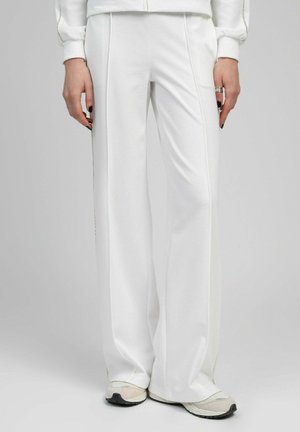 Pantalon blanc à larges jambes porté avec des baskets beiges, mains visibles avec du vernis à ongles noir et des bagues argentées, debout sur un fond uni.