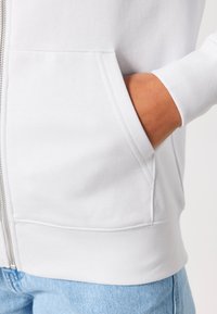 Felpa bianca con zip, dotata di tasche frontali, polsini e vita a coste. Tessuto morbido, design minimalista senza modelli visibili.