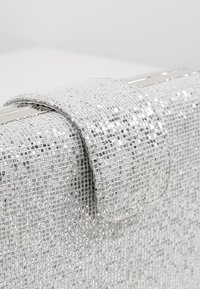 Pochette en métal argenté avec une finition texturée, présentant un rabat repliable et une fermeture en métal lisse. Détails scintillants tout au long.