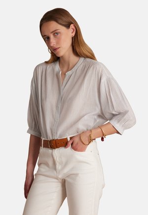 Camicia bianca traspirante e leggera con sottili righe blu, bottoni frontali e vestibilità morbida. Abbinata a jeans bianchi e cintura marrone.