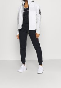 Biała kurtka na suwak z czarnymi akcentami, zestawiona z czarnymi dresami z bocznym logo oraz białymi butami sportowymi. Nowoczesny, stylowy design.