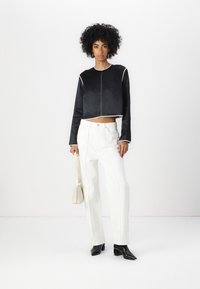 Black cropped jacket with white piping, paired with high-waisted white jeans and black pointed heels. Model holds a small beige handbag.
