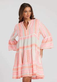 Różowa i biała sukienka w wzory z kołnierzykiem, długimi dzwonowymi rękawami oraz warstwowym dołem z geometrycznymi wzorami w pomarańczowym i różowym kolorze.