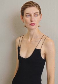 Lühi pruunide juustega naine, kes kannab musta õlata kleiti õhukeste rihmadega, vaatab paremale, seistes lihtsa heleda seina taustal.