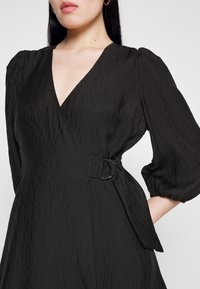 Robe noire enveloppante avec un décolleté en V, en tissu texturé, manches bouffantes et une ceinture latérale avec un accent de boucle circulaire.