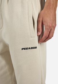Beige joggebukser med en myk tekstur, med sidelommer og svart "PEGADOR" broderi på låret. Nærbilde av stoffet og detaljene.