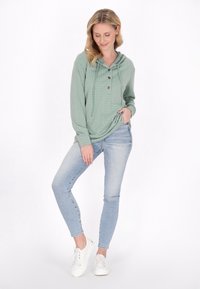 Felpa verde con trama, tasca frontale, tre bottoni in legno, indossata con jeans skinny azzurri e sneakers bianche.