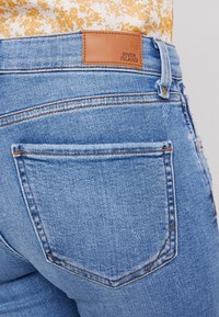 Světle modré džíny s vysokým pasem, s hnědou koženou cedulkou označenou "RIVER ISLAND" a dvěma zadními kapsami s prošíváním.