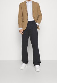 Hnědý blazer přes modro-bíle kostkovanou košili, doplněný námořnickými kalhotami a bílými teniskami. Blazer má dva knoflíky a kapsu.