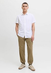 Chemise blanche à manches courtes et à boutons, fabriquée à partir d'un tissu léger, associée à un pantalon kaki fuselé et des baskets beiges avec des accents texturés.