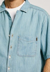 Camisa de mezclilla de manga corta en azul claro, con cuello, bolsillo frontal y cierre de botones. Textura suave con un ligero desgastado y costuras en contraste.