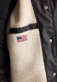Mustale quiltattu takki, jossa on vaaleanbeige tekoshapahuovitus, sivutasku mustalla reunuksella ja pieni Yhdysvaltojen lipun kangastäys.