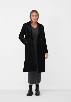 Schwarzer doppelt geknöpfter Mantel mit strukturiertem Muster, großen Revers und Seitentaschen, getragen über einem grauen Kleid und schwarzen Knöchelstiefeln.