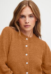 Hnědý, chlupatý pletený cardigan s kulatým výstřihem a pěti dekoračními knoflíky se květinovým vzorem. Měkká textura, volný střih.