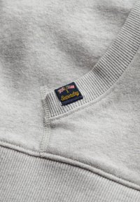 Grijze sweatshirt met ribgebreide halslijn en boorden; voorzien van een geborduurd marineblauw embleem met "Superdry" en vlagaccenten.