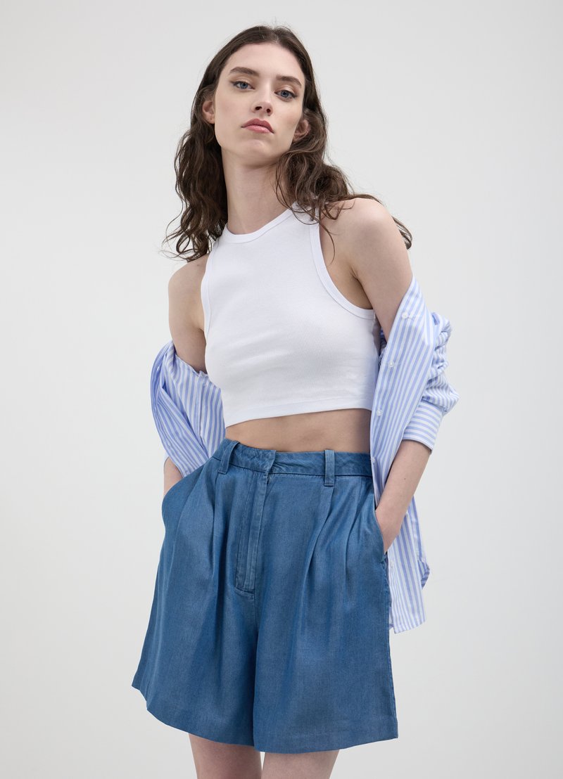 Un haut court côtelé blanc associé à un short en denim plissé taille haute. Une chemise à rayures bleu clair est drapée sur une épaule.