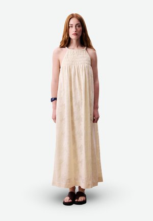 Jeune femme aux longs cheveux roux portant une robe maxi crème sans manches et des sandales noires, se tenant devant un fond blanc uni.